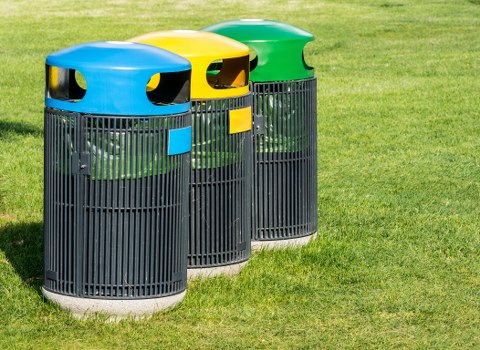 Three cylindrical outdoor rubbish bins positioned on a grassy area, each with a distinct coloured lid in blue, yellow, and green. The bins are made of black plastic with vertical slats forming a ventilated design, and each lid features openings for waste disposal. The blue-lid bin is in the foreground, followed by the yellow and green-lid bins arranged in a slight curve behind it. The background shows a well-maintained lawn, indicating an outdoor setting typical of urban or residential communal spaces in Hackney or nearby areas. The natural lighting suggests daytime conditions. This setup is commonly used for waste and recycling segregation in urban environments, supporting local rubbish collection and clearance services like those offered by Hackney House Clearance.