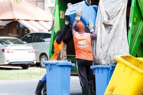 Two waste collection workers wearing orange high-visibility vests and gloves are seen loading a large blue plastic rubbish bin into a green waste collection vehicle on a residential street, with a yellow wheelie bin positioned nearby. One worker is wearing a headscarf, while the other has protective gloves. In the background, there are parked cars, a building with a striped awning, and trees, indicating a suburban area. The scene is well-lit, capturing the process of refuse removal, which is relevant to local rubbish clearance services in Hackney or nearby areas, as represented by Hackney House Clearance's service context.