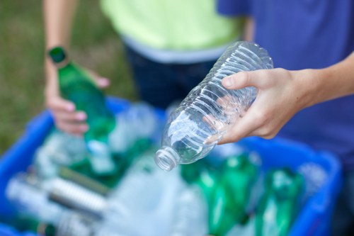 A close-up view of a person's hand holding a clear plastic bottle over a blue recycling bin filled with various green and transparent plastic bottles and containers, set outdoors on a paved surface. In the background, another individual wearing a green shirt and a watch is partially visible, holding additional plastic bottles. The scene depicts a waste sorting or recycling activity, with the focus on plastic waste collection, relevant to rubbish removal services in Hackney or the surrounding London area, as provided by Hackney House Clearance. The environment appears well-lit with natural daylight, emphasizing the clean and orderly collection of recyclable materials prepared for disposal or recycling processing.