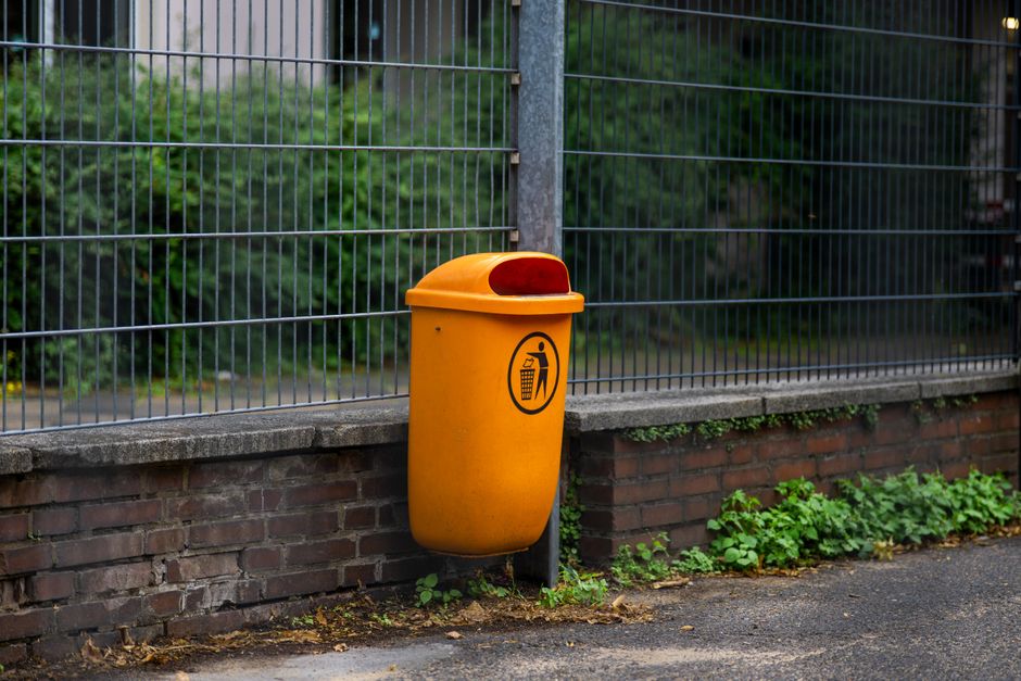 A pile of discarded rubbish on the side of a street, consisting of black plastic trash bags, loose paper and packaging, and an overturned, dirty yellow plastic container. The rubble is resting against