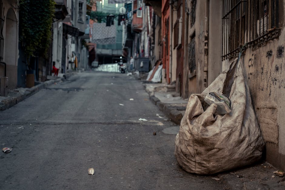 A dirty, off-white fabric rubbish bag filled with waste materials, placed on a narrow, uneven pavement alongside a weathered, graffiti-marked brick wall in an urban alleyway. The bag appears well-used