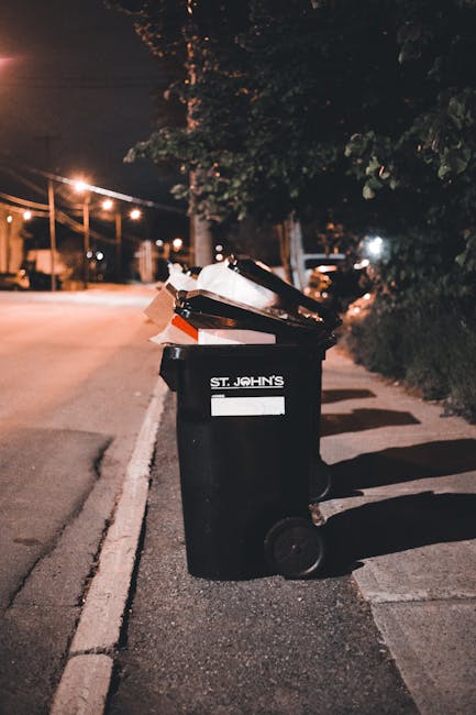A dirty, off-white fabric rubbish bag filled with waste materials, placed on a narrow, uneven pavement alongside a weathered, graffiti-marked brick wall in an urban alleyway. The bag appears well-used